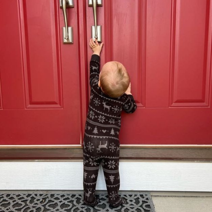 Baby standing at red door wearing The Black Sheep Fam Black Fair Isle Baby Onesie